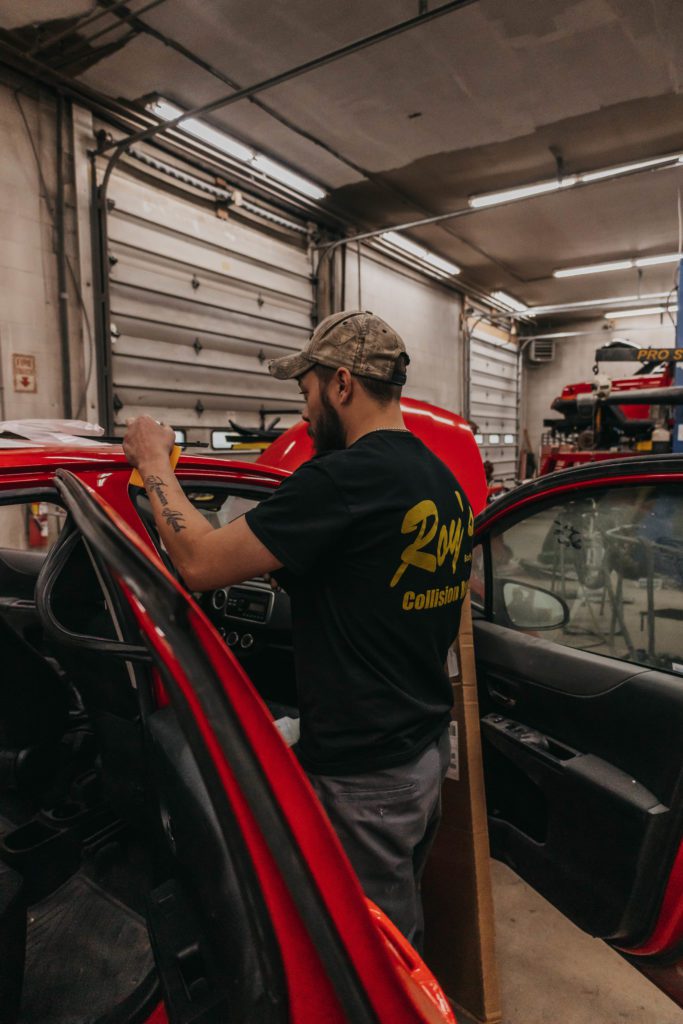 Technician working on a red vehicle inside an auto body repair shop, wearing a shirt with "Roy's Collision" logo, demonstrating hands-on collision repair techniques.