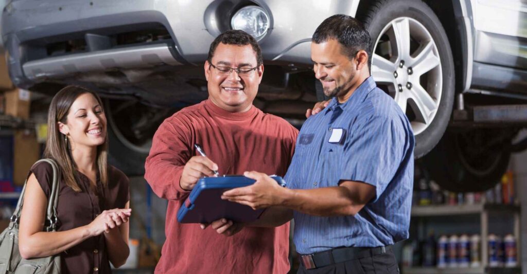 Smiling mechanic in blue shirt holding clipboard as man in red sweater signs, woman in background observing, in auto repair shop setting.
